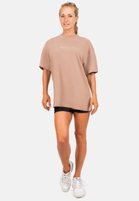 Overgrote taupe T-shirt van katoen met korte mouwen en geborduurd logo. Gecombineerd met zwarte shorts en witte sneakers.