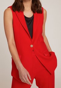 Blazer sans manches rouge avec un bouton unique, coupe ajustée, deux poches avant et une texture de tissu lisse. Assorti avec un haut noir.