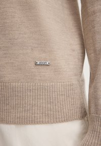 Maglione a maglia beige con orlo a coste; presenta un'etichetta metallica con il logo "JOOP!" attaccata sul davanti. Tessuto morbido, design casual.