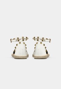 Weiße Leder-Espadrilles mit goldenen Pyramidennieten, Knöchelriemen und einer gewebten Jutesohle; offene Rückseite und Seiten für Belüftung.