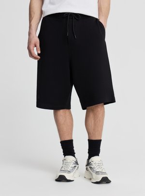 Mężczyzna w czarnych szortach do kolan ze sznurkiem, czarnych skarpetkach, białych i szarych sneakersach oraz białej koszuli, stojący na szarym tle.