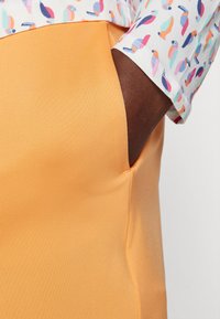 Pantalones de tela naranja con una textura suave y bolsillo lateral; la cinturilla se combina con una camisa de estampado ligero que presenta coloridos pájaros.