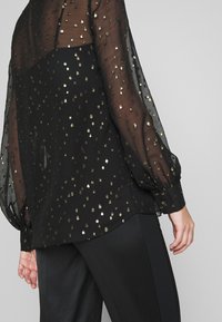 Blouse noire transparente à pois dorés, manches longues bouffantes, associée à un pantalon noir sur un fond gris uni.