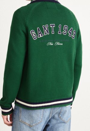 Personne portant un pull vert tricoté avec le texte "GANT 1949" dans le dos et un jean bleu, vue de dos.