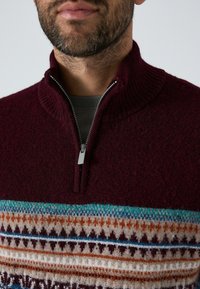 Bordeaux ritsweater met geribbelde kraag. Heeft een gemusterde band in tinten blauw, oranje en crème over de borst. Zachte textuur.