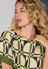 Mustrilineaalne tipp, millel on hele beež alus, musta, rohelise ja kollase geomeetrilise kujundusega, lõdvestunud lõige, lai kaelus ja lühikesed varrukad.