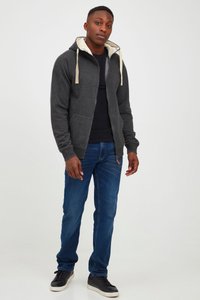 Junger Mann steht, trägt eine dunkelgraue Kapuzenjacke mit Reißverschluss, schwarzes Shirt, blaue Jeans und schwarze Sneaker mit weißen Sohlen vor einem schlichten Hintergrund.