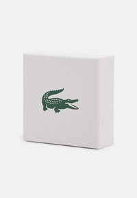 Weiße Box mit einer strukturierten Oberfläche, die ein grünes Krokodil-Logo zeigt. Die Box hat eine rechteckige Form und schräg verlaufende Kanten.