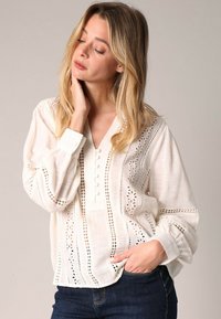 Blouse en coton blanc avec un col en V, détail de boutons et des motifs découpés complexes le long des manches et sur le devant. Portée avec un jean noir.