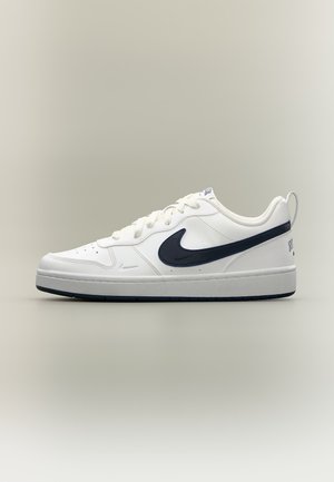 Zapatilla baja blanca de Nike con logotipo swoosh negro, cierre de cordones y suela de goma sobre un fondo claro y liso.
