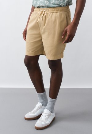 Άνδρας που φοράει μπεζ σορτς με κορδόνι, γκρι ριμπ κάλτσες, λευκά αθλητικά παπούτσια και πράσινο πουκάμισο με σχέδια, στέκεται σε γκρι δάπεδο.