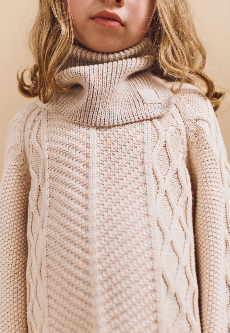 Barn med langt blondt hår iført en cremefarvet tyk strikket sweater med høj rullekrave.