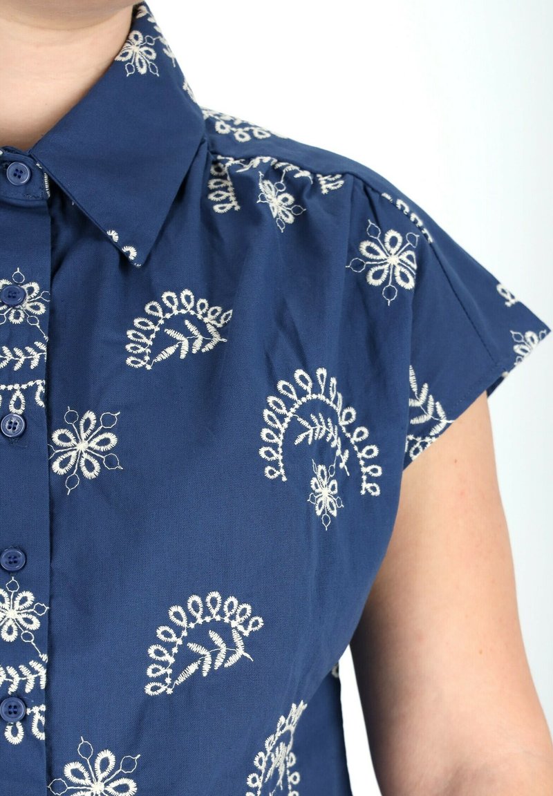 Camicetta blu navy a maniche corte, con motivi ricamati floreali bianchi e bottoni frontali. Tessuto leggero con una consistenza liscia.