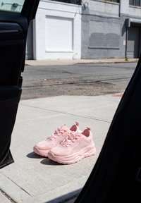 Paire de baskets roses sur un trottoir en béton, vue à travers une porte de voiture ouverte avec un bâtiment gris et une rue en arrière-plan.