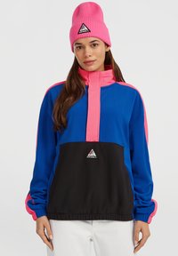 Kvinde iført en blå, sort og pink jakke med en pink hue, stående mod en ensartet lys grå baggrund.