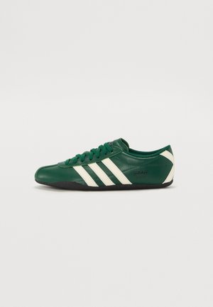 Groene leren sneaker met witte strepen, ronde neus, vetersluiting, zwarte zool en "adidas TOKYO" merknaam aan de zijkant.