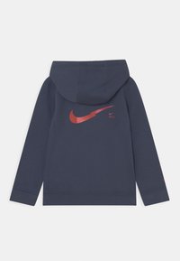 Námořnická mikina s červeným logem Nike swoosh na zadní straně, žebrovanými manžetami a stažitelnou kapucí pro nastavitelné přizpůsobení. Měkká textilie.