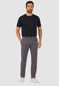 Pantaloni chino grigi con una vestibilità slim e tasche frontali, abbinati a una t-shirt nera aderente. Il modello indossa sneakers bianche e si trova di fronte a uno sfondo neutro.