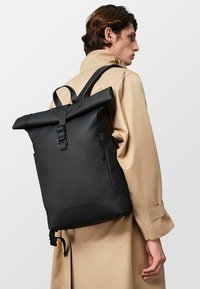Sac à dos noir à rabat fabriqué en matériau durable, doté de bretelles réglables, d'une poche zippée à l'avant et d'un logo en relief sur le bas de l'avant.