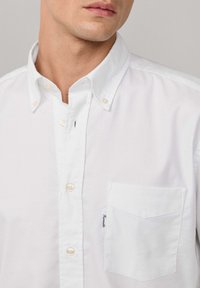 Camisa blanca de botones hecha de algodón con un cuello clásico, botones de perlas y un bolsillo en el pecho izquierdo que presenta una etiqueta. Textura suave, diseño minimalista.