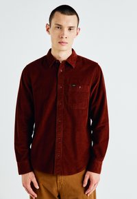 Camisa de pana marrón con cierre de botones en la parte delantera, mangas largas y un bolsillo en el pecho. La tela tiene un acabado texturizado. Diseño simple, sin patrones.
