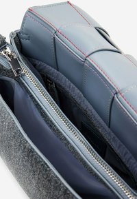 Wnętrze torby z szarego materiału i niebieskiej skóry z srebrnymi zamkami błyskawicznymi, pokazujące dwie przegrody, wyłożone ciemnym materiałem z czerwonymi akcentami szwów.