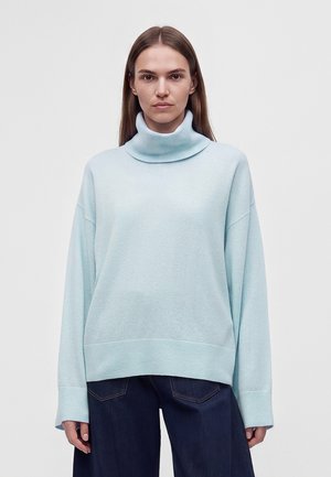 Femme aux cheveux bruns et lisses portant un pull à col roulé bleu clair et un jean bleu foncé, debout devant un fond clair uni.