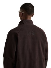 Personne aux cheveux courts portant une veste polaire marron foncé avec "ATW" brodé en haut du dos, vue de dos, porte également des lunettes et une petite boucle d’oreille anneau.