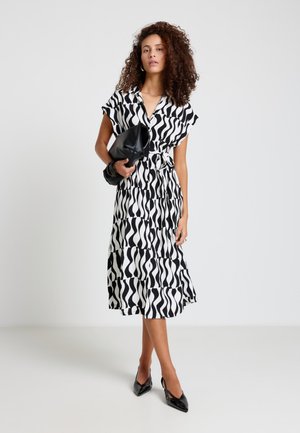 Femme aux cheveux bouclés portant une robe à motif ondulé noir et blanc, tenant une pochette noire, debout avec les jambes croisées en talons noirs.