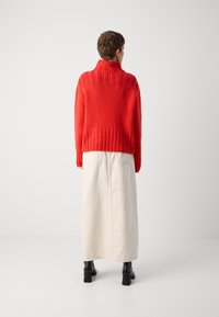 Roter gestrickter Pullover mit hohem Kragen, kombiniert mit einem hellbeigen Maxirock. Schwarze Stiefeletten runden das Outfit ab.