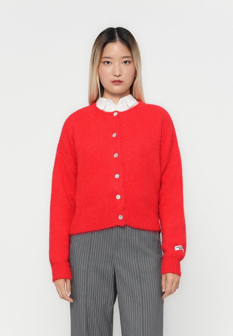 Cardigã vermelho de malha com decote redondo, seis botões prateados e punhos canelados. Usado sobre uma camisa branca de colarinho e combinado com calças cinzas risca de giz.