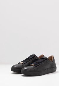 Para czarnych skórzanych sneakersów niskich z czarnymi sznurówkami i beżowym wykończeniem wewnątrz na białej powierzchni z szarym tłem.