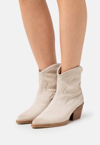 Béžové semišové krátké kozačky se špičatými špičkami, složenými horními částmi a ozdobným šitím. Naskládané dřevěné podpatky přidávají nenápadnou výšku.