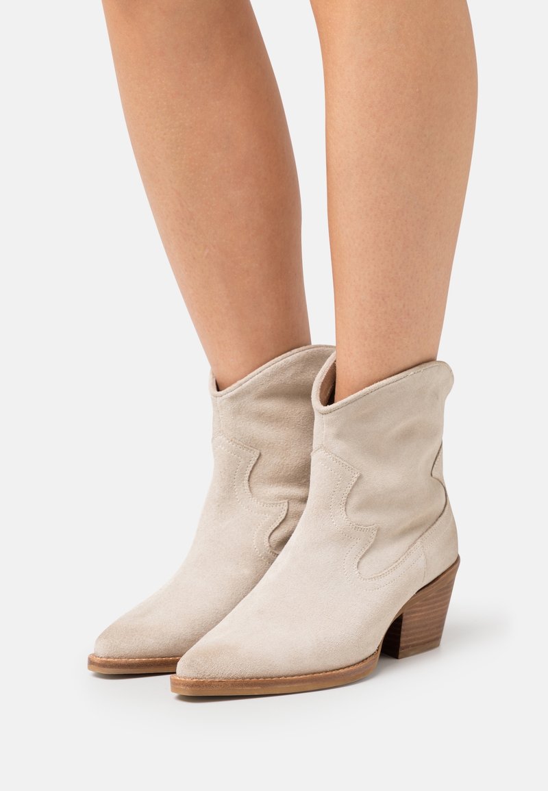 Béžové semišové krátké kozačky se špičatými špičkami, složenými horními částmi a ozdobným šitím. Naskládané dřevěné podpatky přidávají nenápadnou výšku.