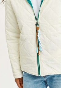 Prešívaná bunda v off-white farbe s tyrkysovými akcentmi, obsahujúca predný zips so dekoratívnym modro-hnedým ťahadlom a dlhými rukávmi.