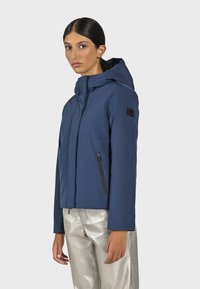 Mörkblå jacka med huva och dragkedja fram, med svarta dragkedjeelement och en logotyp-lapp på ärmen; slätt, lätt material.
