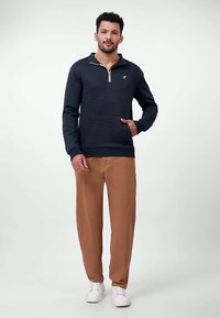 Tumši zils pulovers ar rāvējslēdzēja apkakli un zelta emblēmu. Kombinēts ar brūnām biksēm. Modelis stāv baltās sporta kurpēs uz gaiša fona.