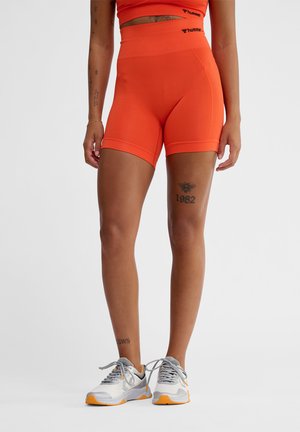 Person iført lyse orange shorts med høj talje og matchende top, med synlige tatoveringer på benene og grå og orange atletiske sko.