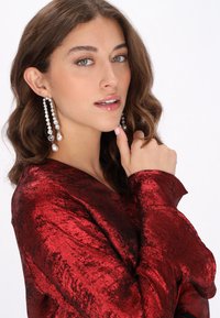 Rød velourtop med tekstureret finish, lange ærmer og dyb halsudskæring. Lange, hængende øreringe med krystaller og perledekorationer.