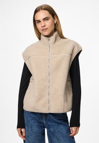 Vestă bej din fleece cu guler înalt, fermoar frontal și decupaje la umeri, purtată peste o bluză neagră cu mâneci lungi, asortată cu blugi albaștri.