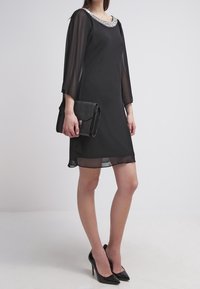 Robe noire avec des manches transparentes et un col en perles, assortie à une pochette noire et des escarpins pointus, illustrant un design simple et élégant.