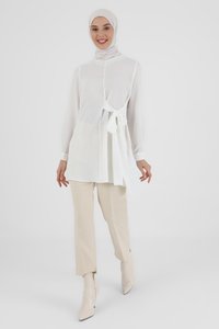 White long-sleeve top with a tie detail at the waist, layered over beige trousers and paired with pointed toe beige ankle boots. Smooth fabric.