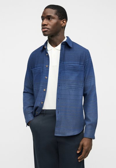 Άνδρας που φοράει μπλε ναυτικό πουκάμισο με κουμπιά πάνω από λευκό πόλο και σκούρο παντελόνι, στέκεται με το αριστερό χέρι στην τσέπη μπροστά σε απλό φόντο.