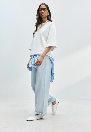 Woman wearing oversized white shirt, light blue wide-leg jeans, white shoes, blue striped shirt tied around waist, and sunglasses standing on white floor.