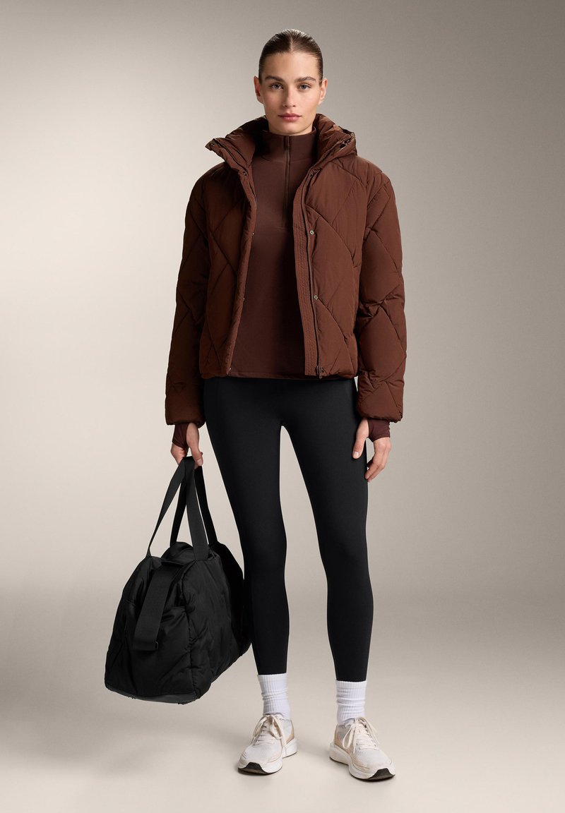 Femme en doudoune marron, haut marron, leggings noirs, baskets blanches, tenant un grand sac de sport noir, debout devant un fond uni.