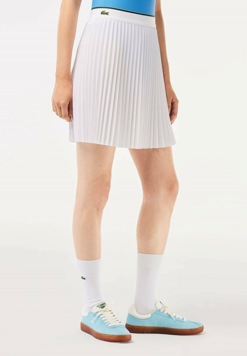 Falda blanca con pliegues, cintura ancha y logo de Lacoste. Combinada con zapatillas azul claro y calcetines blancos. Textura suave, diseño de longitud media.