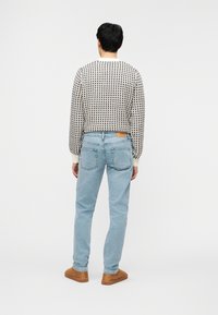 Άνδρας με πουλόβερ σε άσπρο και μαύρο καρό, ανοιχτό μπλε τζιν και καφέ σουέτ παπούτσια, στέκεται με θέα σε απλό λευκό φόντο.