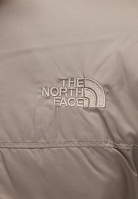 Țesătură bej cu logo-ul brodat "The North Face" în gri, însoțit de un simbol distinctiv de munte curbat. Textură netedă, finisaj durabil.
