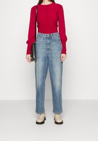 Rotes langärmeliges Rippstrick-Oberteil, kombiniert mit hochgeschnittenen hellblauen Jeans; das Modell hält eine schwarze Schultertasche und trägt beigefarbene Loafers.