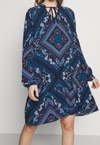 Långärmad klänning i marinblått med intrikata paisleymönster, som har geometriska former i rött och turkos, samt en nyckelhålshalsringning med snörning.